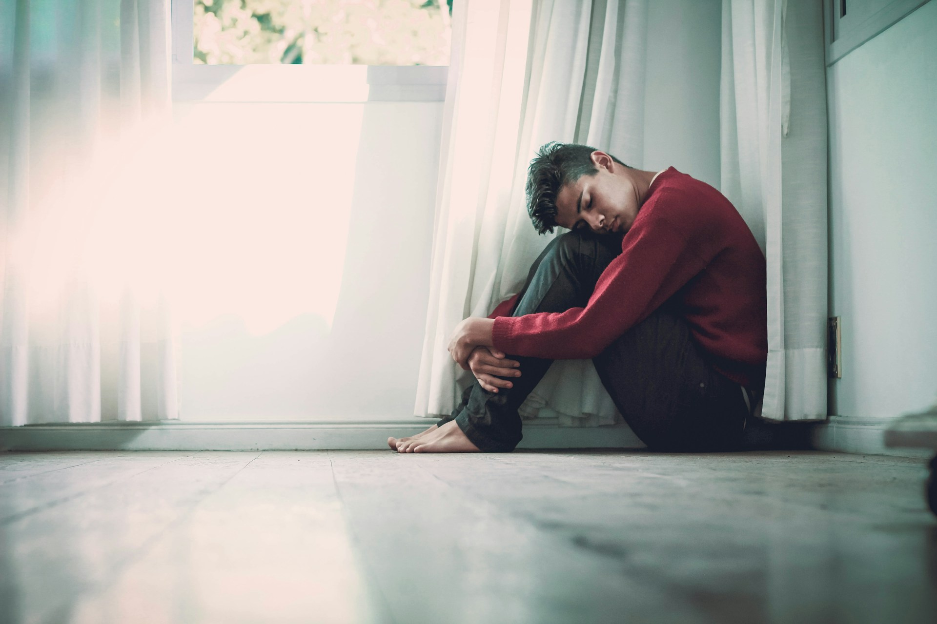 A man sits on the floor, wrapped in their arms, against a wall, surrounded by soft natural light filtering through curtains.