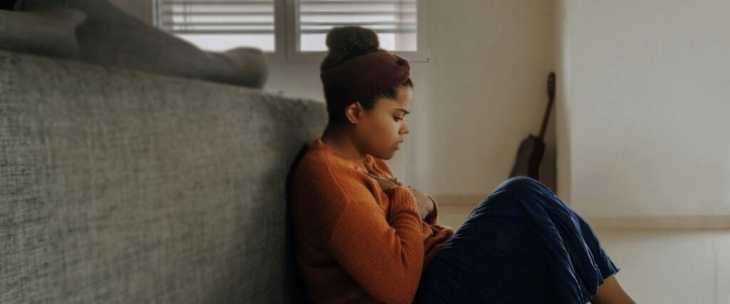 A woman sits on the floor beside a sofa, wearing an orange sweater and blue skirt, with a calm atmosphere and soft natural lighting.