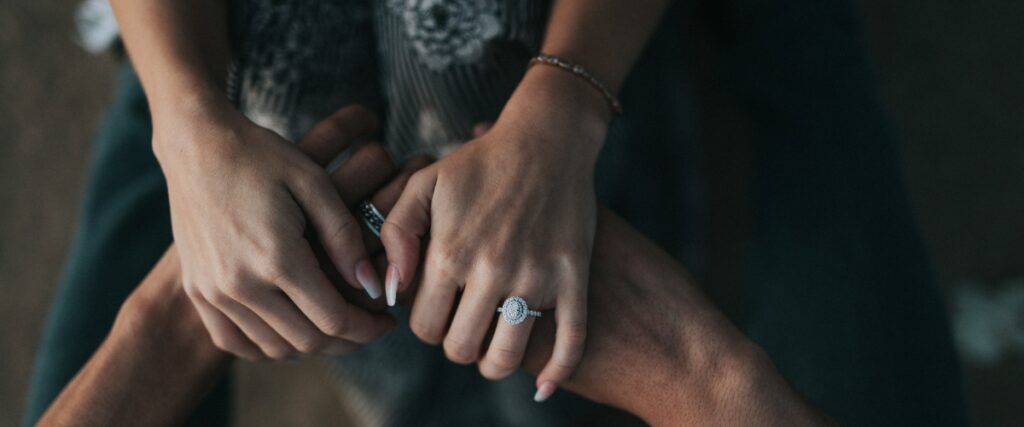 Close-up of two hands intertwined, showcasing a sparkling engagement ring on one hand amid a warm, intimate setting.