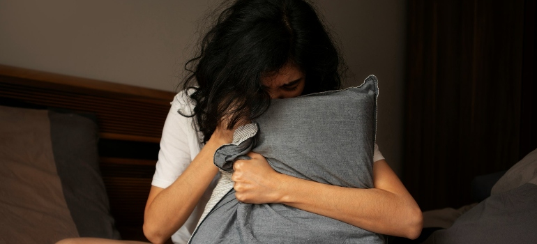 A woman with dark curly hair sits on a bed, hugging a gray pillow tightly, with a distressed expression on her face.