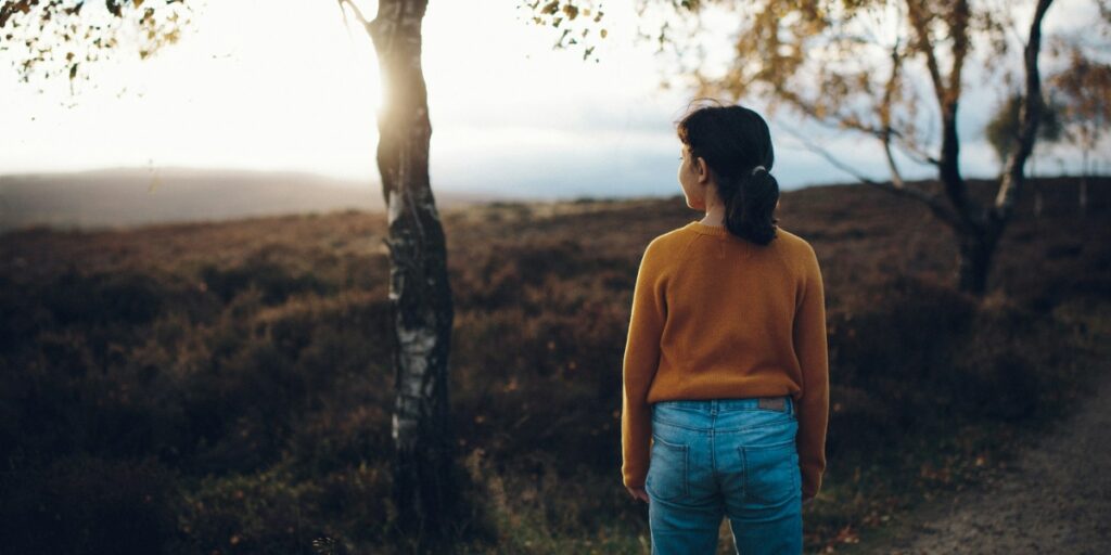 A young woman in a mustard sweater and blue jeans stands by a tree, gazing thoughtfully into a serene landscape at sunset.
