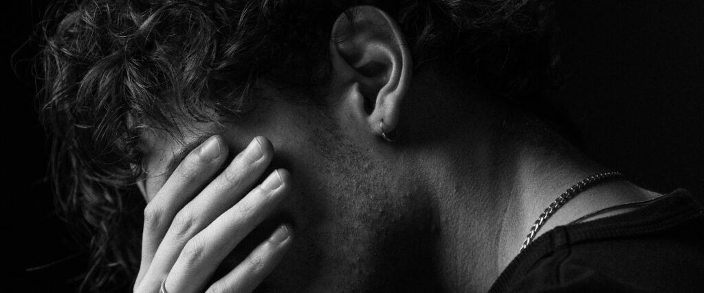 A close-up of a man with curly hair, his head bowed and hand covering his face in a moment of distress, in soft black and white lighting.