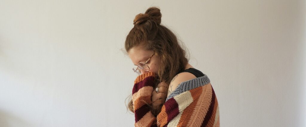 A person with wavy, brown hair styled in a bun, wearing a colorful, knitted sweater, leans slightly forward against a plain white wall.