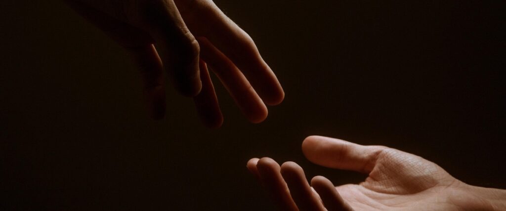 Two hands reach toward each other against a dark background, symbolizing connection and humanity.