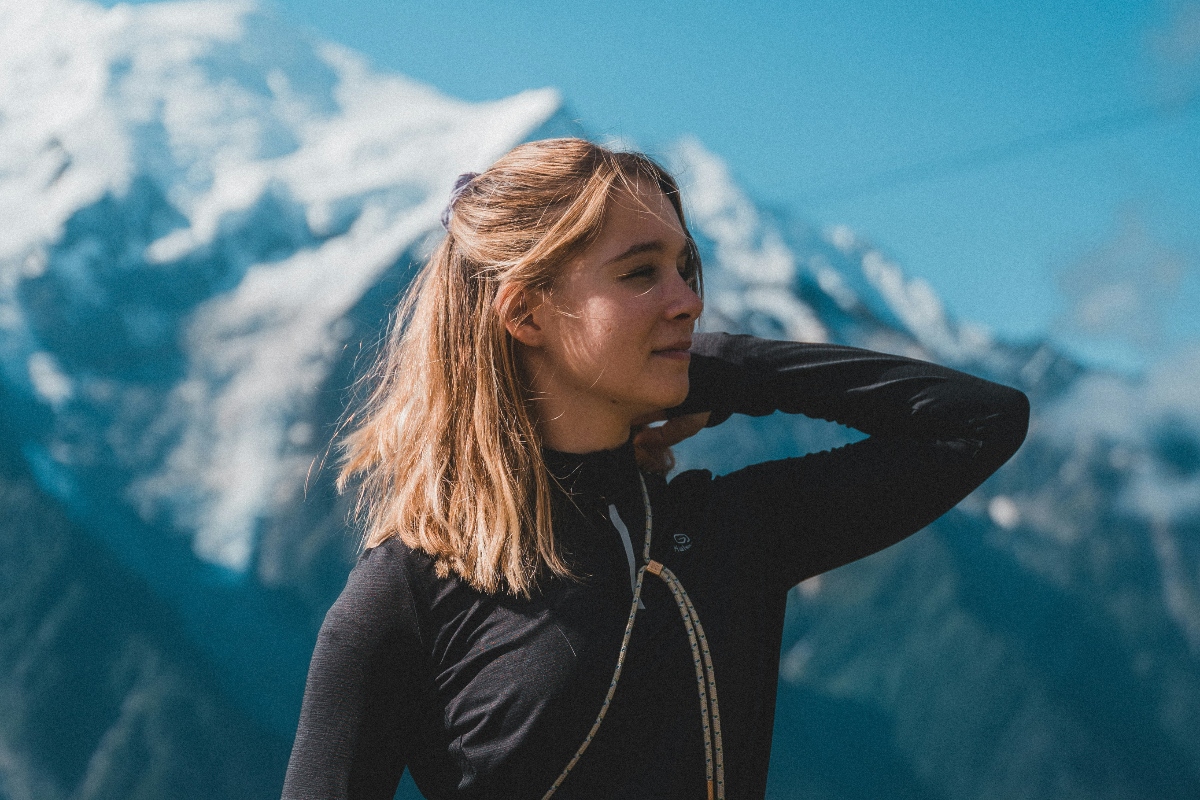 A person in a black long-sleeve top stands against a backdrop of majestic snow-capped mountains under a clear blue sky.
