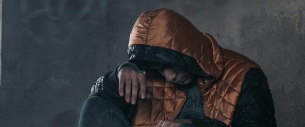 A young man in a hooded jacket sits against a wall, looking down, conveying a sense of sadness and introspection, considering Virginia fentanyl addiction treatment.