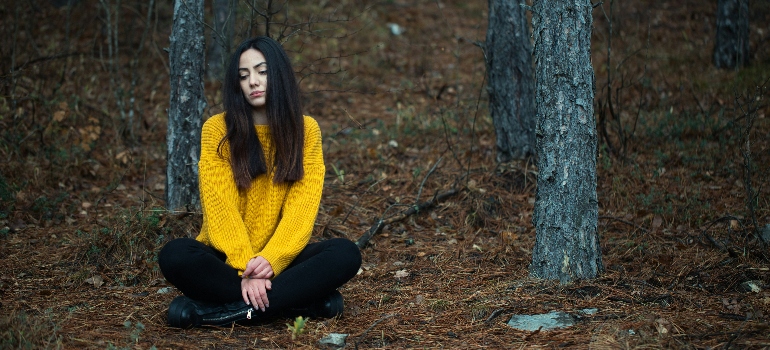 A person sits cross-legged on the forest floor, wearing a bright yellow sweater and black pants, surrounded by tall trees and fallen leaves.
