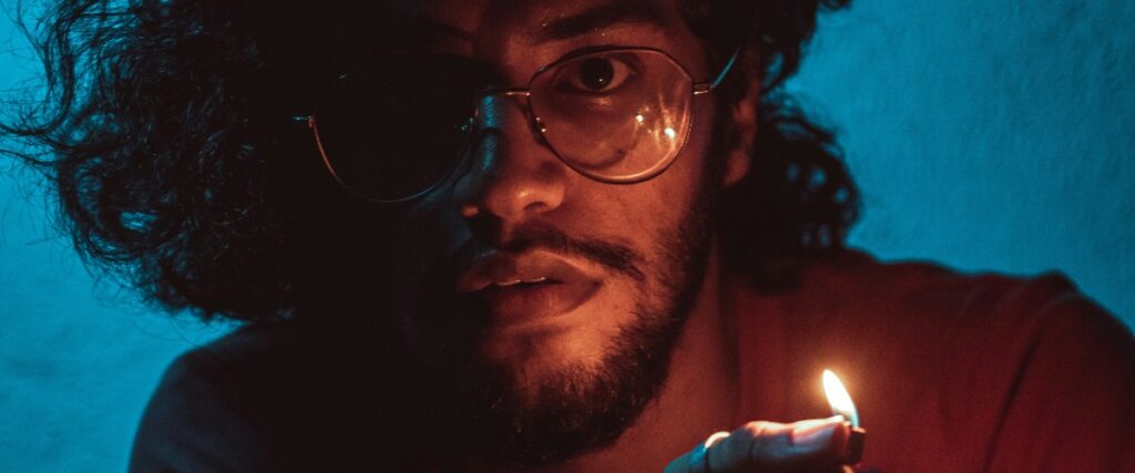A person holding a lit match, illuminated against a dark blue background, surrounded by curls of hair and shadows.