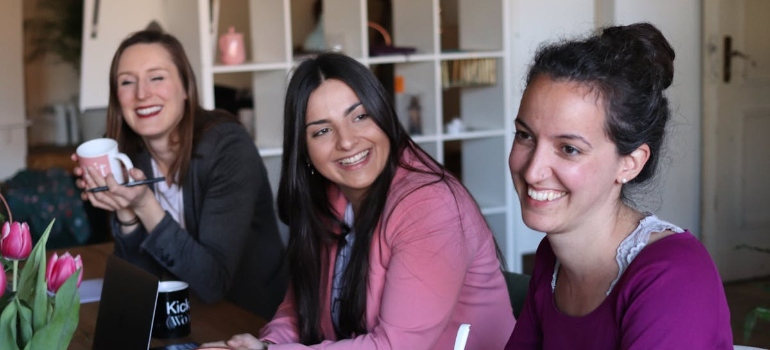 Three women smile and converse in a warm, casual office setting. Two hold mugs, while pink tulips on the table add a touch of color and freshness.