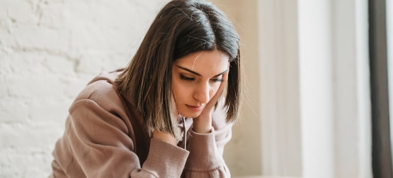 A woman in a cozy sweatshirt sits with her head lowered, conveying a sense of introspection and sadness in a softly lit room.