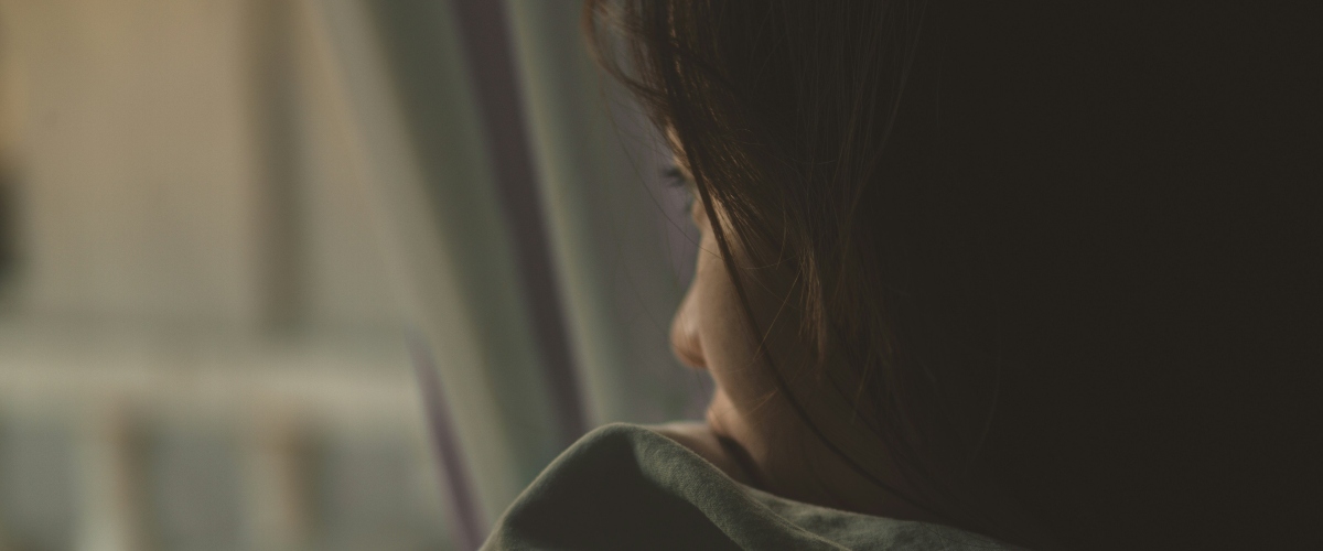 A person sits near a window, draped in soft fabric, with warm sunlight filtering through sheer curtains, creating a serene atmosphere.