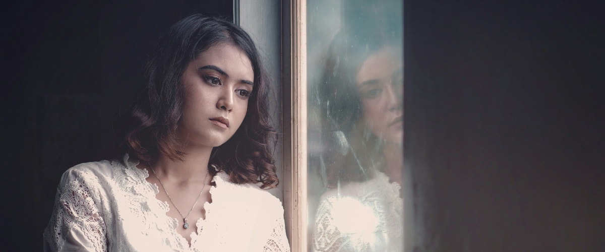 A woman stands by a window, wearing a white lace blouse, with soft curls framing her face and a reflective glass in the background.