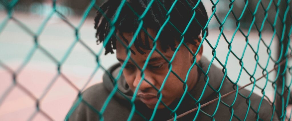 A person with dreadlocks leans against a chain-link fence, set against a softly blurred background of a sports court.