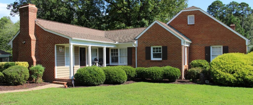 A charming brick house with a gabled roof, surrounded by manicured shrubs and a grassy lawn, under a clear blue sky.