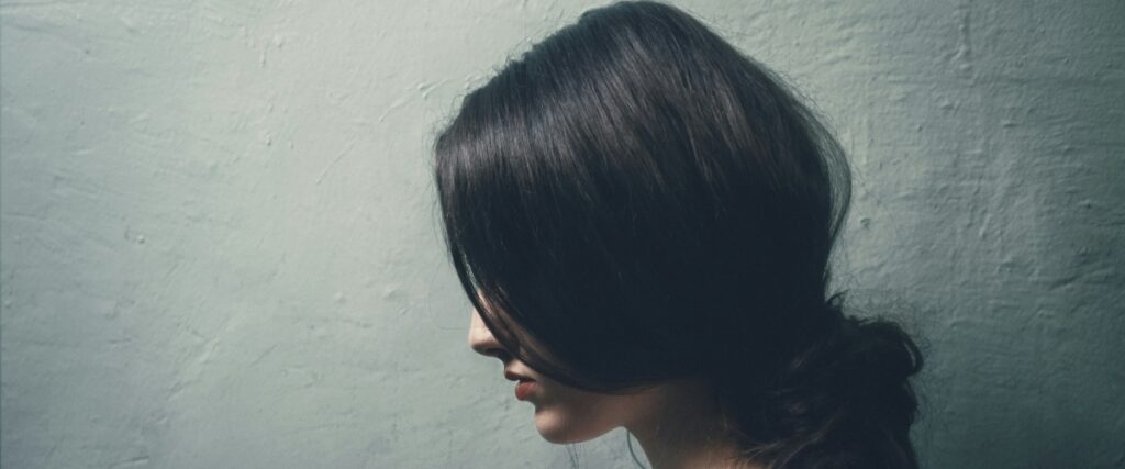 A profile view of long, dark hair cascading over a shoulder against a textured, light gray wall.